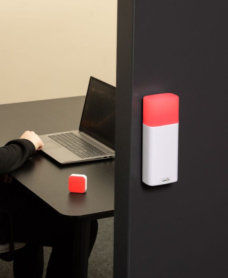 Luxafor Switch Pro 2 on a meeting room wall glowing red, with a Cube physical switch on the desk inside showing the same red status