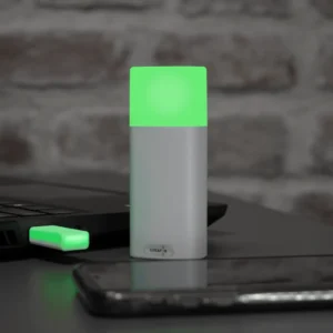 Luxafor Bluetooth Pro wireless busy light glowing green on a dark desk next to a laptop and smartphone, with USB Bluetooth dongle connected