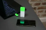 Luxafor Bluetooth Pro wireless busy light glowing green on a dark desk next to a laptop and smartphone, with USB Bluetooth dongle connected