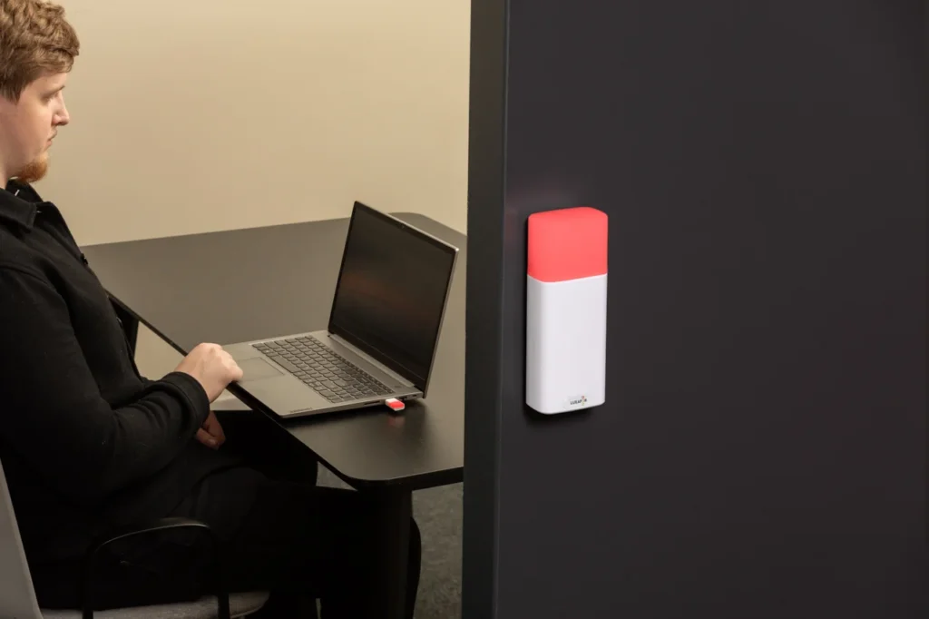 Luxafor Bluetooth Pro wireless busy light glowing red, wall-mounted on a dark office partition, with a man working on a laptop visible in the background and a USB dongle in the laptop