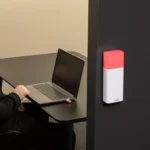 Luxafor Bluetooth Pro wireless busy light glowing red, wall-mounted on a dark office partition, with a man working on a laptop visible in the background and a USB dongle in the laptop