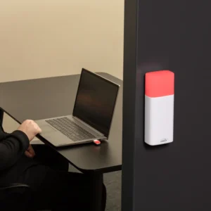 Luxafor Bluetooth Pro wireless busy light glowing red, wall-mounted on a dark office partition, with a man working on a laptop visible in the background and a USB dongle in the laptop